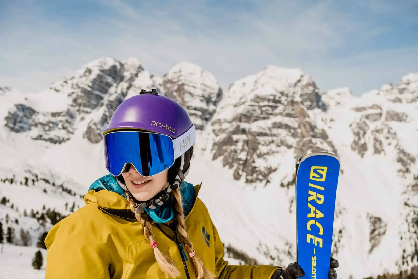 Woman with blonde hair standing on a mountain top holding a pair of skis