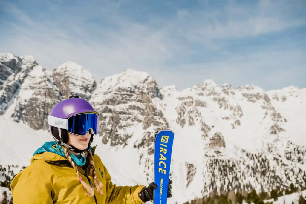Blonde female skier wearing a yellow jacket holding a pair of blue Salomon skis and wearing photochromic ski goggles from Messy Weekend