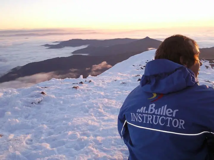 Image of a male Mt Buller ski instructor sitting on a mountain top at sunset wearing a ski instructor jacket 