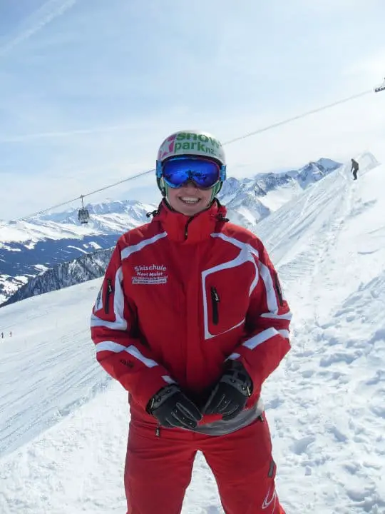 A photo of a female snowboard instructor wearing a read ski suit, white helmet and blue ski goggles