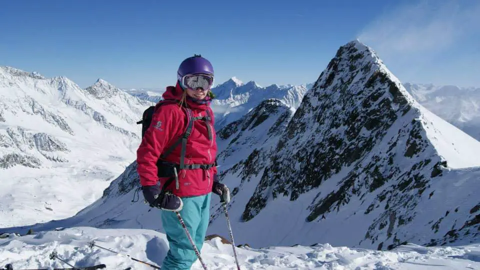 Photo of a female skier in a pink jacket standing on a mountain top on a clear blue day with tall mountain peaks around her.